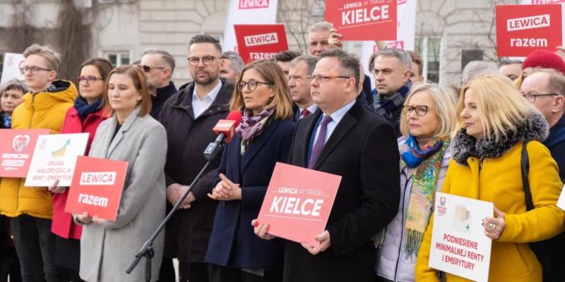 Na zdjęciu widoczna jest grupa osób biorących udział w konferencji prasowej, trzymających transparenty z hasłami politycznymi, w tym „LEWICA w trasie KIELCE” i „LEWICA razem”.