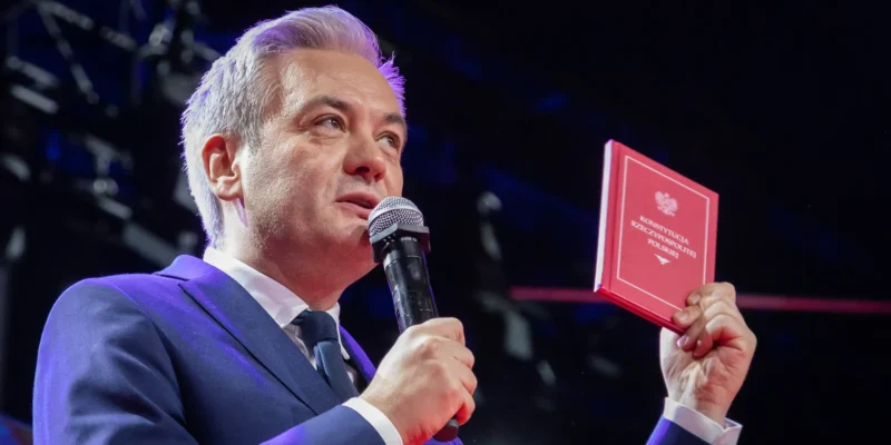 Mężczyzna w granatowym garniturze przemawia na scenie, trzymając w jednej ręce mikrofon, a w drugiej czerwony podręcznik Konstytucji RP.