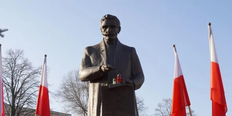 Pomnik postaci stojącej w garniturze w otoczeniu flagi Polski i drzew, na tle jasnego nieba.