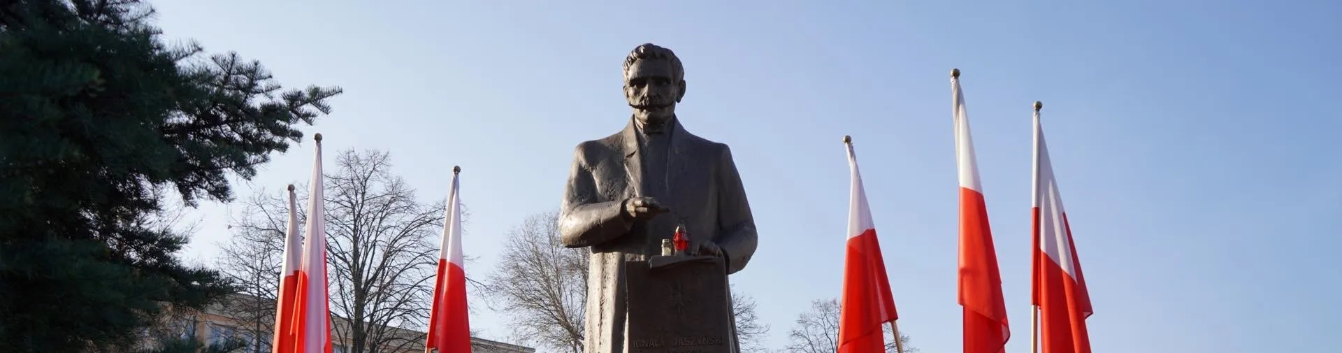 Pomnik postaci stojącej w garniturze w otoczeniu flagi Polski i drzew, na tle jasnego nieba.