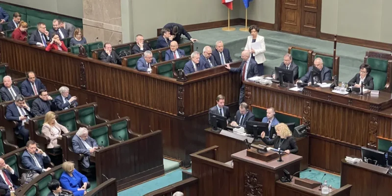 Na zdjęciu widać posiedzenie parlamentu, z przedstawicielami różnych ugrupowań politycznych siedzącymi w drewnianych ławach.