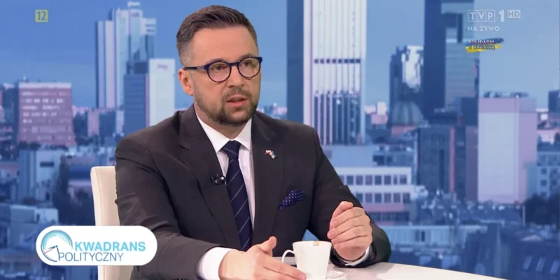 Ujęcie z programu Kwadrans Polityczny: mężczyzna w garniturze rozmawia przy kawie w studiu telewizyjnym, na tle panoramy miasta.