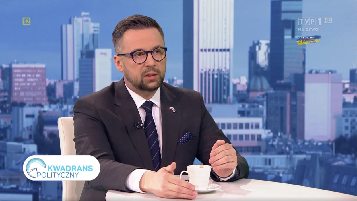 Ujęcie z programu Kwadrans Polityczny: mężczyzna w garniturze rozmawia przy kawie w studiu telewizyjnym, na tle panoramy miasta.