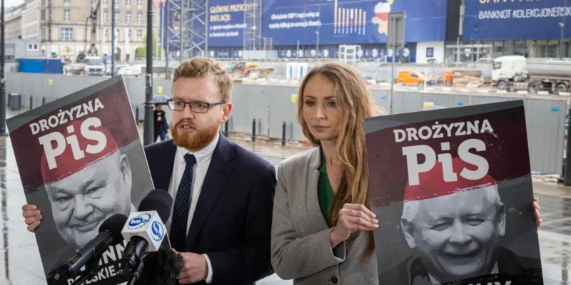 Dwóch mężczyzn w garniturach stoi na placu budowy, trzyma plakaty z napisem PiS i wizerunkiem uśmiechniętego dziecka podczas konferencji z mikrofonami.