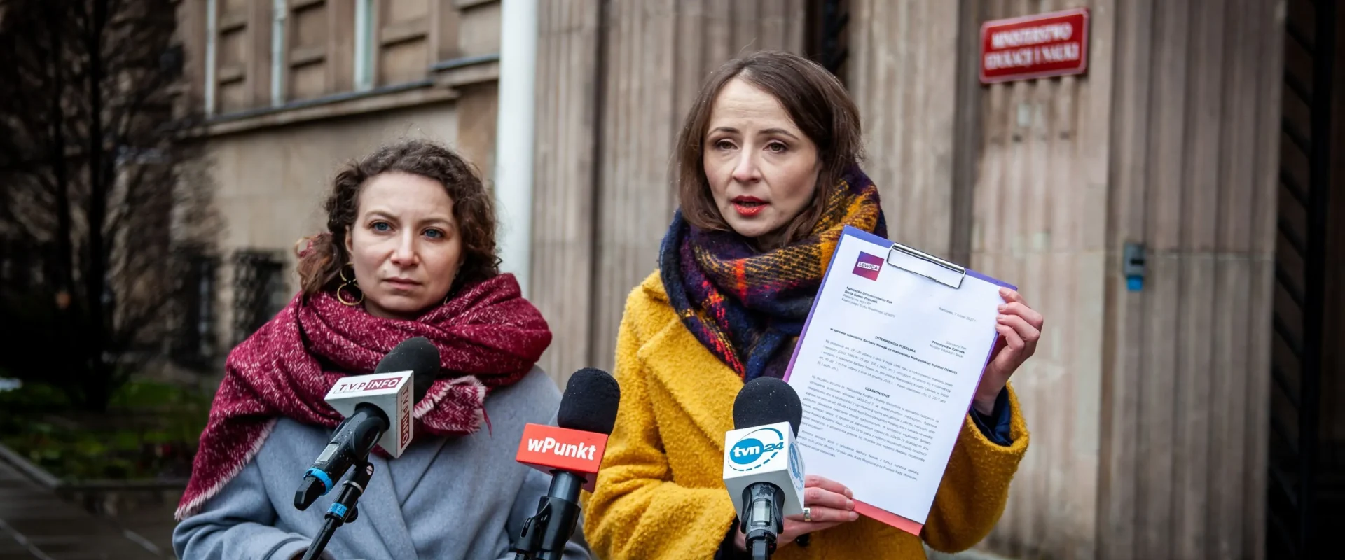 Dwie kobiety w zimowych płaszczach stoją przed mikrofonami podczas konferencji prasowej na zewnątrz budynku urzędu; jedna z nich trzyma kartkę z dokumentem.
