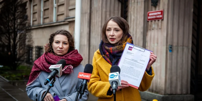 Dwie kobiety w zimowych płaszczach stoją przed mikrofonami podczas konferencji prasowej na zewnątrz budynku urzędu; jedna z nich trzyma kartkę z dokumentem.