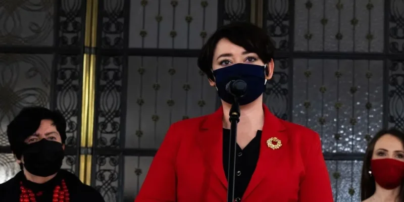 Polityczka w czerwonej marynarce przemawia do mikrofonu podczas konferencji prasowej, w tle dwie osoby w maseczkach.