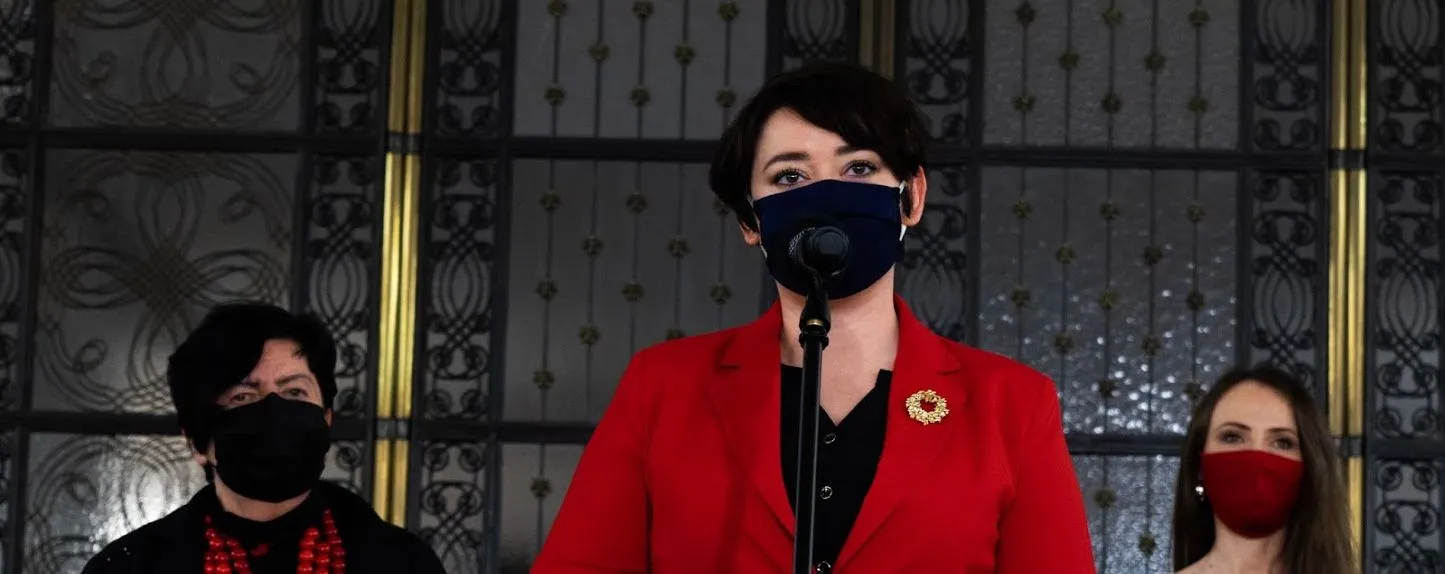 Polityczka w czerwonej marynarce przemawia do mikrofonu podczas konferencji prasowej, w tle dwie osoby w maseczkach.