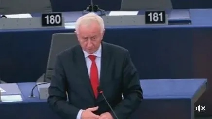 Mężczyzna w garniturze i czerwonym krawacie przemawia przy mównicy na sali obrad z numerowanymi miejscami.