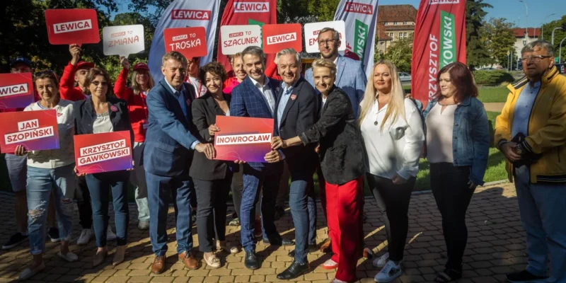 Grupa osób na zewnątrz trzyma plakaty i flagi Lewicy z hasłami promującymi profilaktykę w ochronie zdrowia.