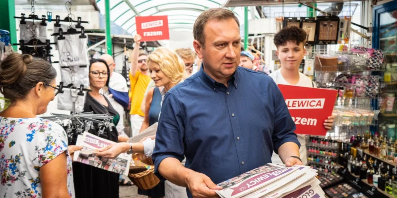 Na zdjęciu widać mężczyznę z gazetami w rękach, przechodzącego przez targ, wokół niego są ludzie z transparentami wspierającymi ruch Lewicy.