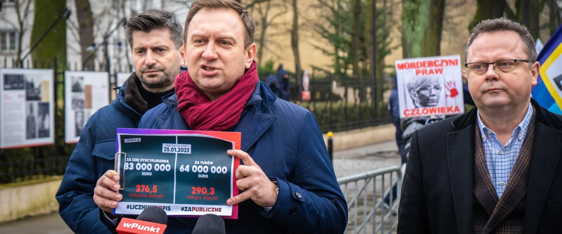 Na zdjęciu znajduje się mężczyzna w granatowym płaszczu i czerwonym szaliku, trzymający plakaty z danymi finansowymi, podczas publicznej manifestacji z hasłami o prawach człowieka w tle.