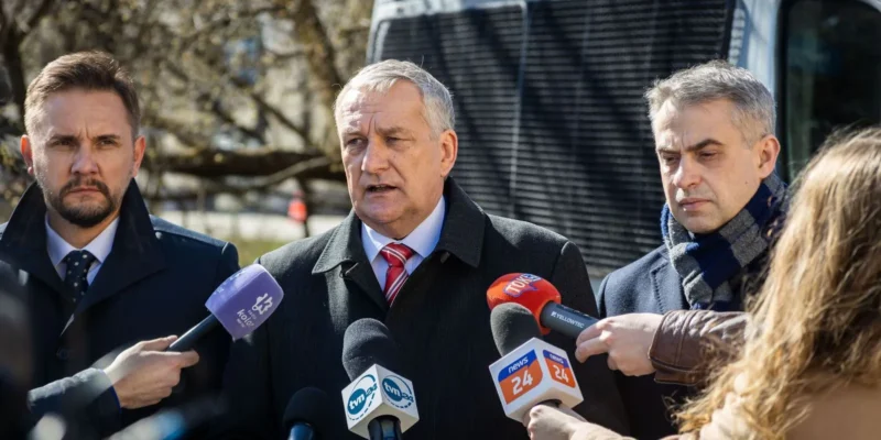 Trzech mężczyzn w garniturach udziela konferencji prasowej, stojąc przed mikrofonami reporterów, a w tle widać część pojazdu policyjnego.