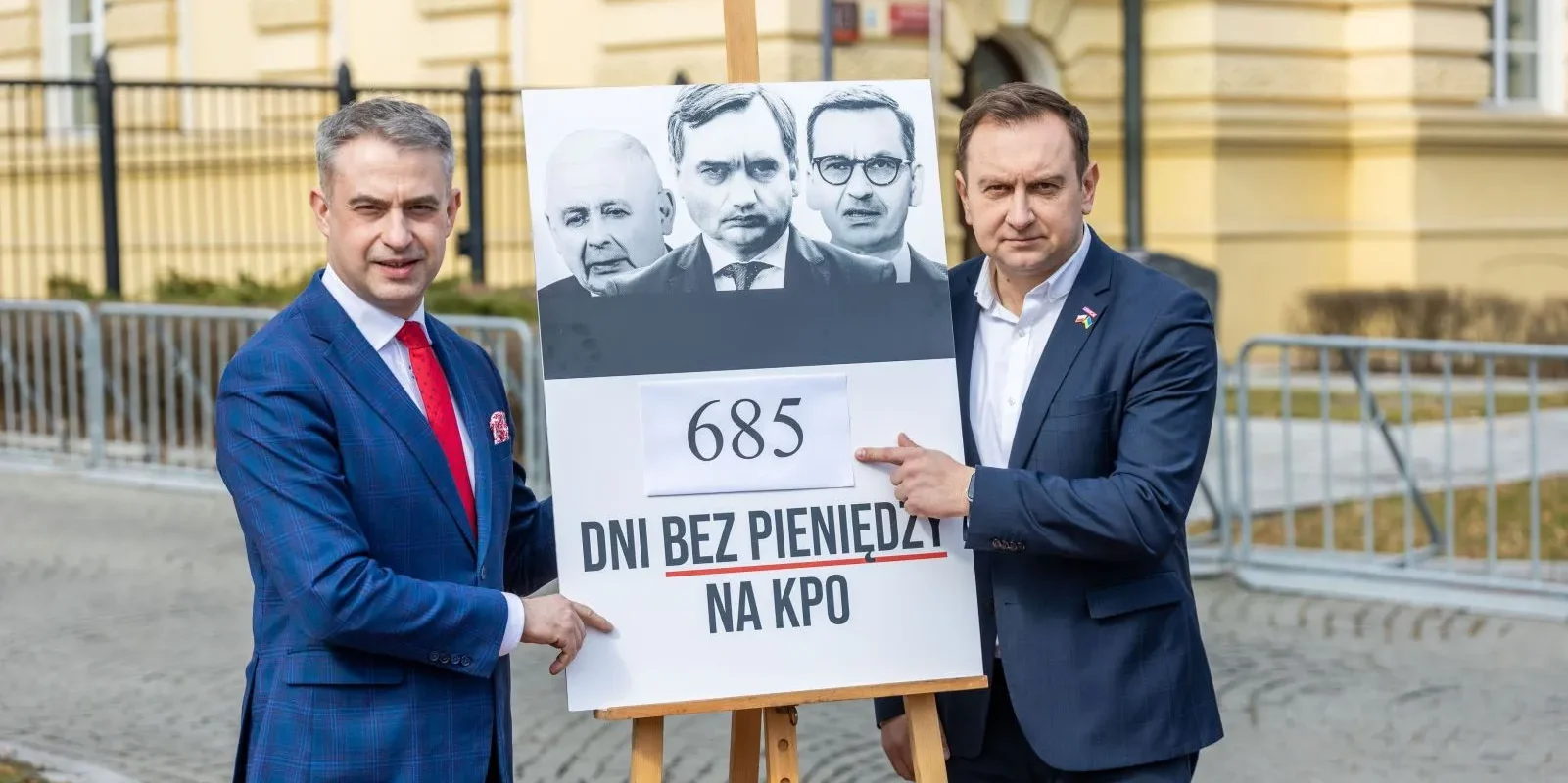 Dwóch mężczyzn w garniturach stoi obok plakatu z napisem „685 dni bez pieniędzy na KPO” i podobiznami polityków.