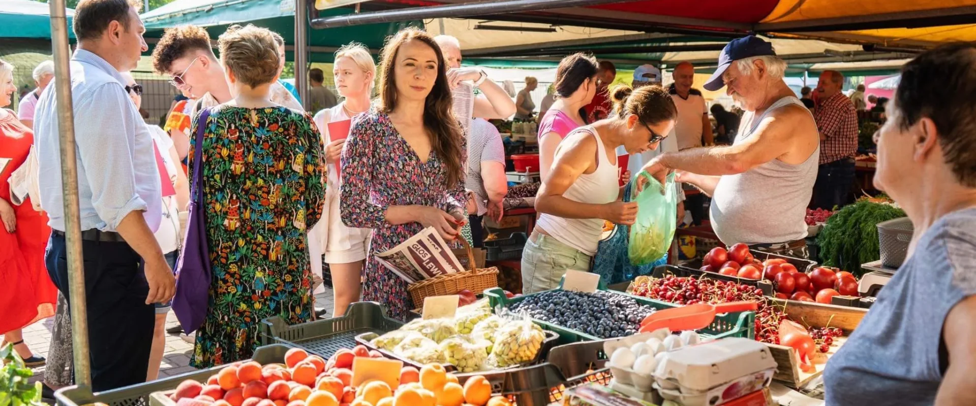 Grupa ludzi na targu pod kolorowymi namiotami wybiera świeże owoce i warzywa, a w centrum stoi kobieta w kwiecistej sukience trzymająca gazetę.