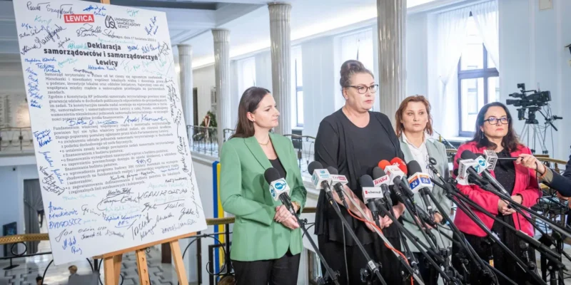 Kobiety stojące przy mikrofonach podczas konferencji prasowej, obok dużej planszy z podpisami i tytułem „Deklaracja samorządowców i samorządowców Lewicy”.