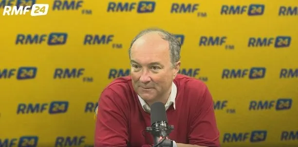 Mężczyzna w czerwonym swetrze siedzi przed mikrofonem w radiowym studiu na tle żółtego baneru RMF24.