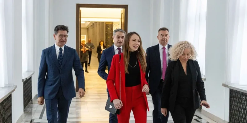 Grupa osób w garniturach spaceruje po eleganckim, jasnym korytarzu; na pierwszym planie kobieta ubrana w czerwony garnitur prowadzi grupę.