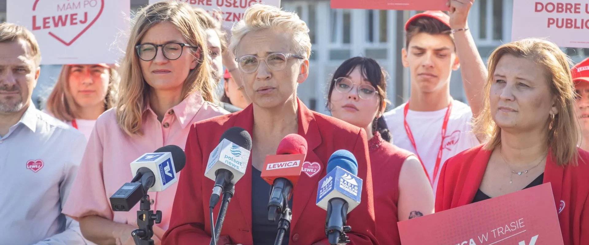 Kobieta w czerwonym żakiecie przemawia na konferencji prasowej przed mikrofonami, otoczona zwolennikami z plakatami Lewicy w kontekście postulatu świeckiej ochrony zdrowia.