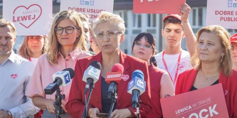 Kobieta w czerwonym żakiecie przemawia na konferencji prasowej przed mikrofonami, otoczona zwolennikami z plakatami Lewicy w kontekście postulatu świeckiej ochrony zdrowia.