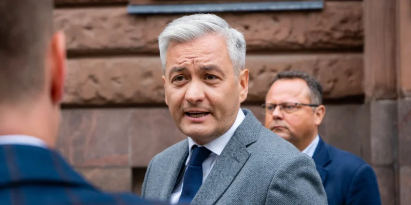 Mężczyzna w szarym garniturze i granatowym krawacie rozmawia z innymi na zewnątrz, na tle kamiennej fasady budynku.