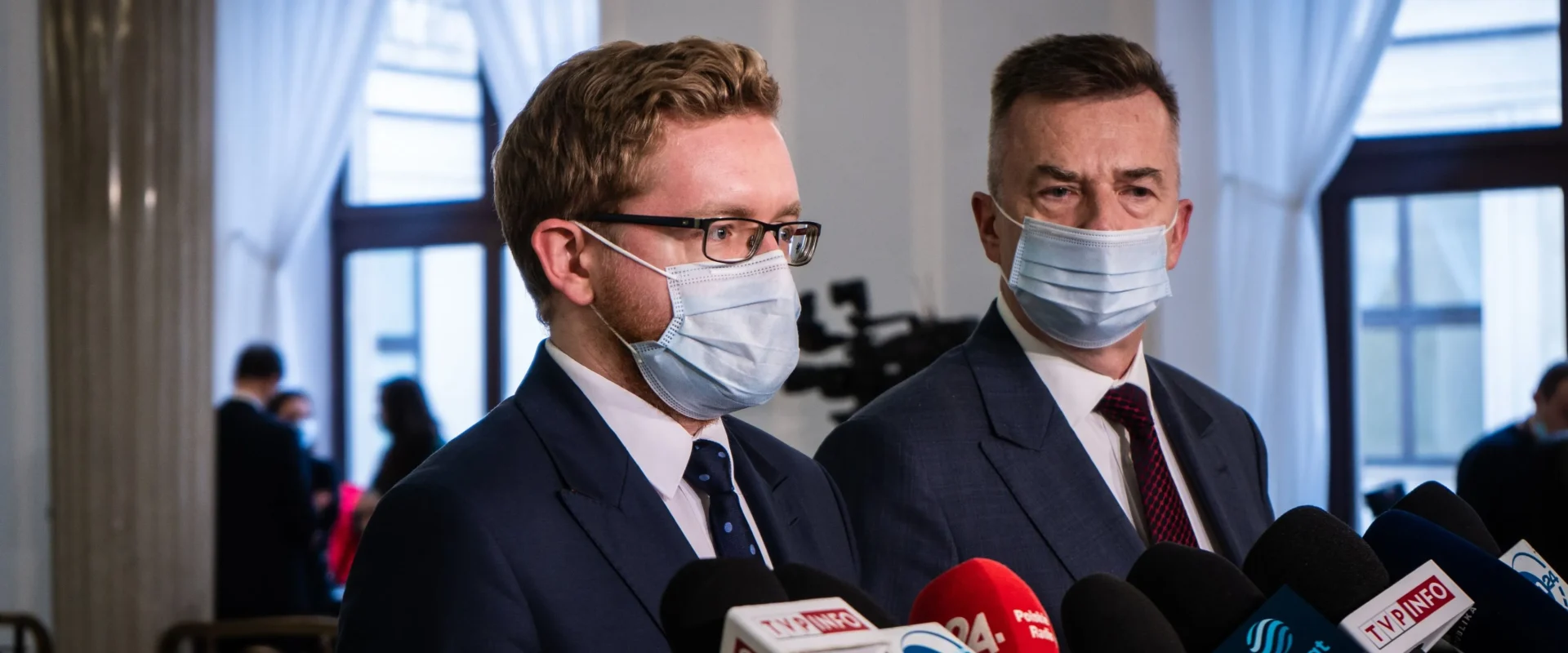 Dwóch mężczyzn w garniturach i maseczkach udziela konferencji prasowej, stojąc przed zestawem mikrofonów dziennikarskich.