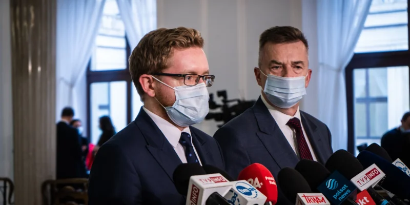 Dwóch mężczyzn w garniturach i maseczkach udziela konferencji prasowej, stojąc przed zestawem mikrofonów dziennikarskich.