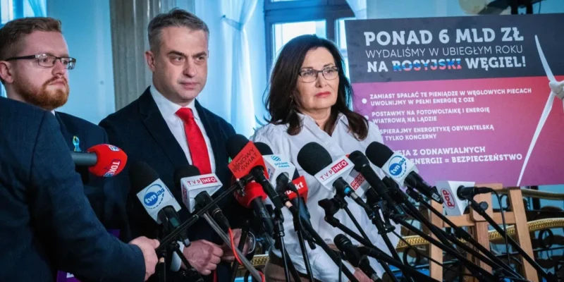 Konferencja prasowa z kilkoma osobami stojącymi przed licznymi mikrofonami, w tle plakat promujący bezpieczeństwo energetyczne i odchodzenie od węgla.