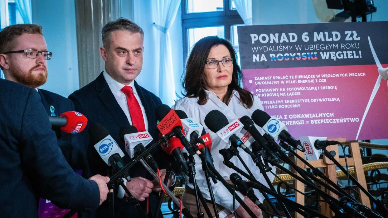 Konferencja prasowa z kilkoma osobami stojącymi przed licznymi mikrofonami, w tle plakat promujący bezpieczeństwo energetyczne i odchodzenie od węgla.