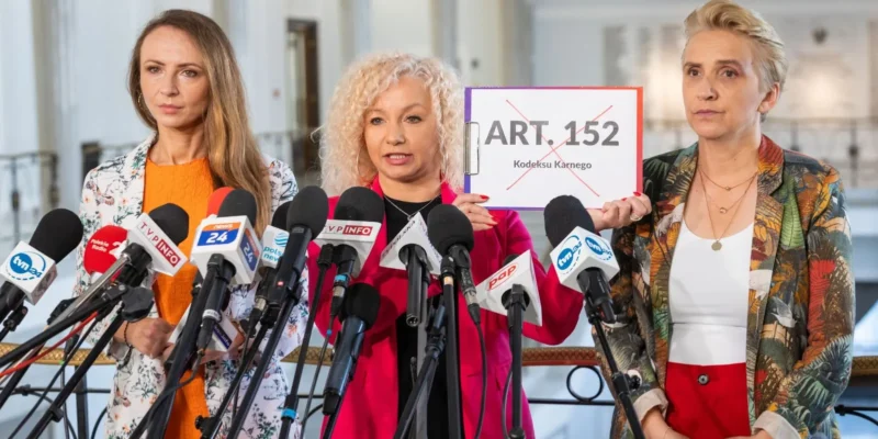 Trzy kobiety stojące na konferencji prasowej za licznymi mikrofonami, aśrodkowa trzyma kartę z napisem „ART. 152 Kodeksu Karnego” z czerwonymi przekreśleniami.