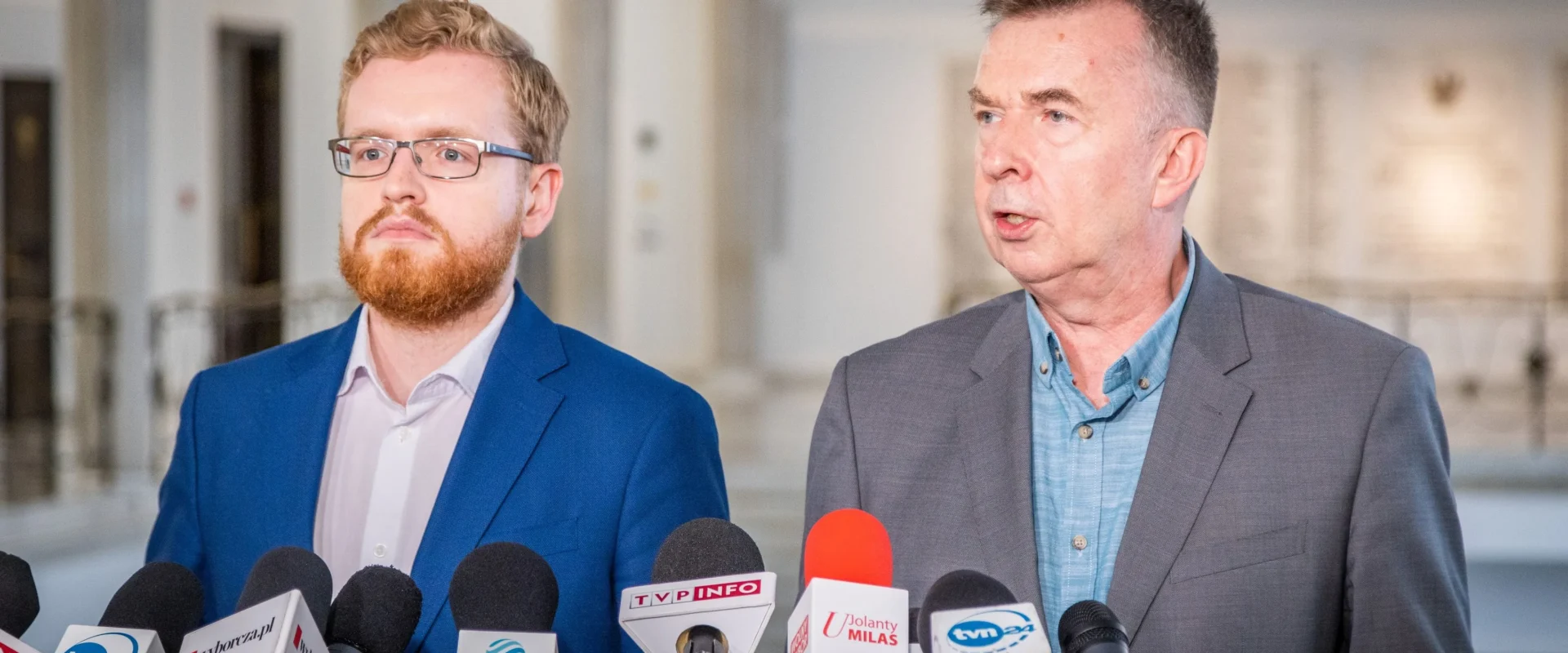 Dwóch mężczyzn w garniturach na konferencji prasowej dotyczącej rozbudowy elektrowni w Ostrołęce, stojących przy licznych mikrofonach mediów.