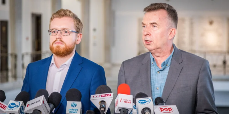 Dwóch mężczyzn w garniturach na konferencji prasowej dotyczącej rozbudowy elektrowni w Ostrołęce, stojących przy licznych mikrofonach mediów.