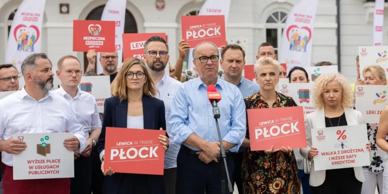 Grupa polityków Lewicy na demonstracji z czerwonymi plakatami, a w centrum mężczyzna przemawia do mikrofonu.