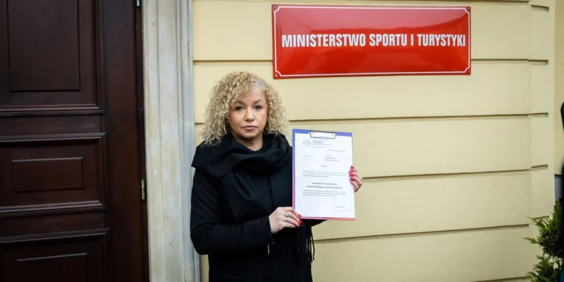 Kobieta o kręconych blond włosach w czarnym płaszu stoi przed budynkiem z tablicą MINISTERSTWO SPORTU I TURYSTYKI i trzyma w dłoni dokument na teczce.