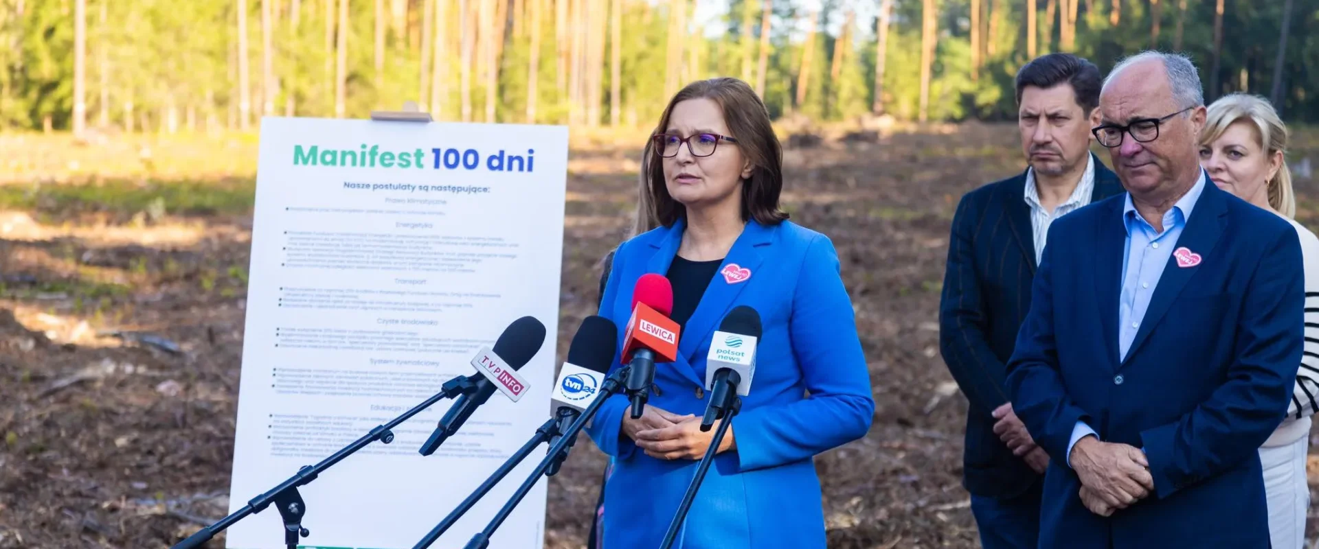 Kobieta w niebieskim garniturze przemawia do mediów podczas konferencji w lesie, stojąc obok tablicy z napisem Manifest 100 dni i kilku mikrofonów.