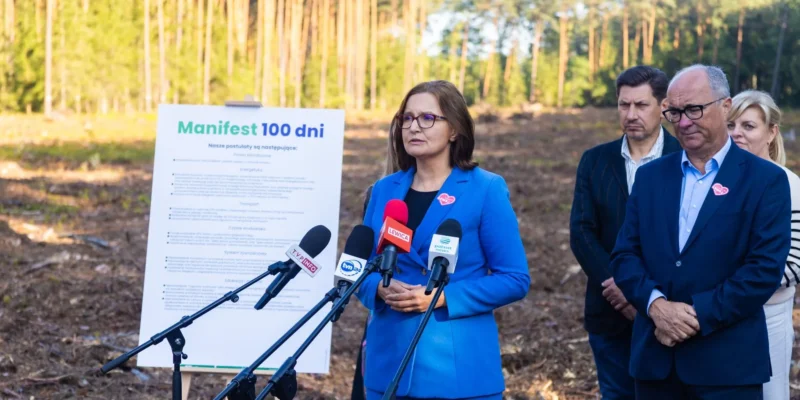 Kobieta w niebieskim garniturze przemawia do mediów podczas konferencji w lesie, stojąc obok tablicy z napisem Manifest 100 dni i kilku mikrofonów.