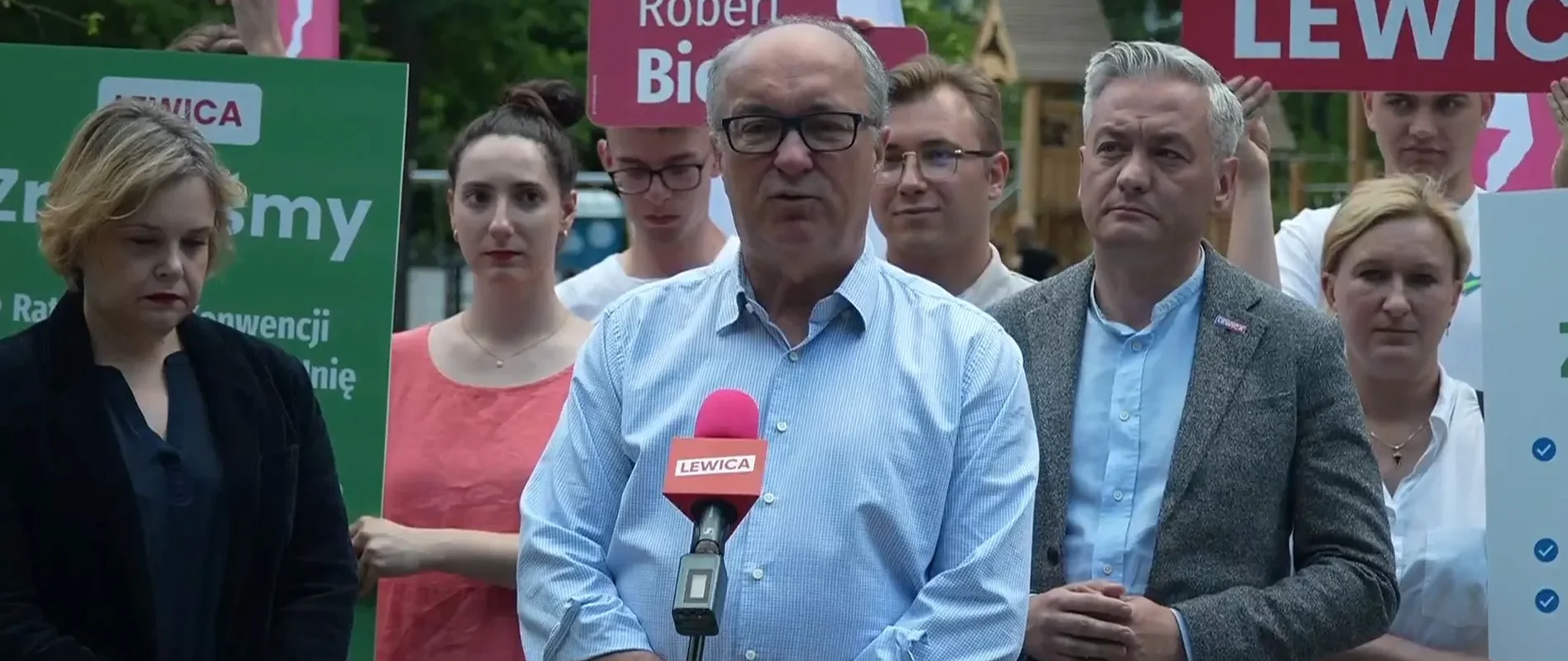 Grupa osób na konferencji prasowej Lewicy; na pierwszym planie mężczyzna w niebieskiej koszuli mówi do mikrofonu z napisem Lewica, w tle stoją inni uczestnicy i plakaty.