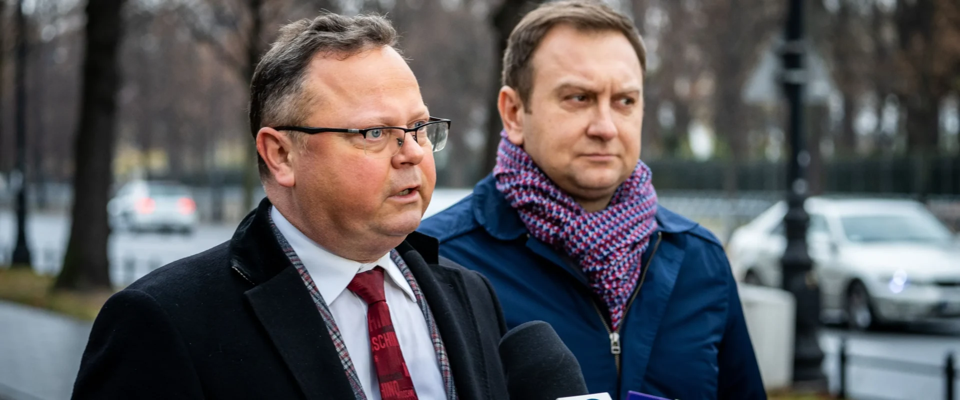 Dwaj mężczyźni, jeden w czarnym płaszczu z krawatem, a drugi w niebieskiej kurtce z kolorowym szalikiem, przemawiają do mediów na ulicy.