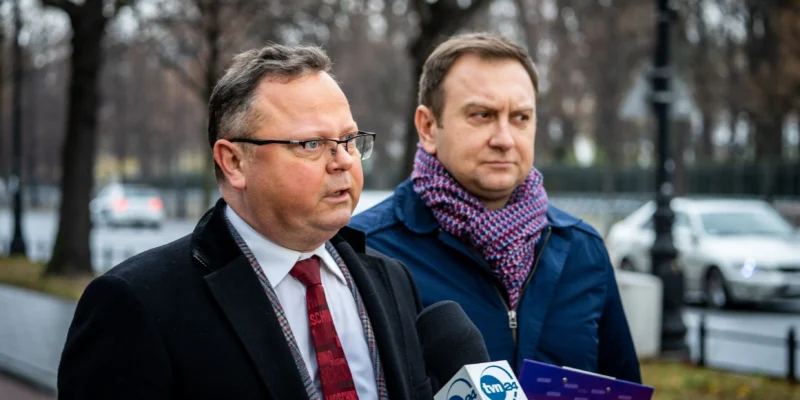 Dwaj mężczyźni, jeden w czarnym płaszczu z krawatem, a drugi w niebieskiej kurtce z kolorowym szalikiem, przemawiają do mediów na ulicy.