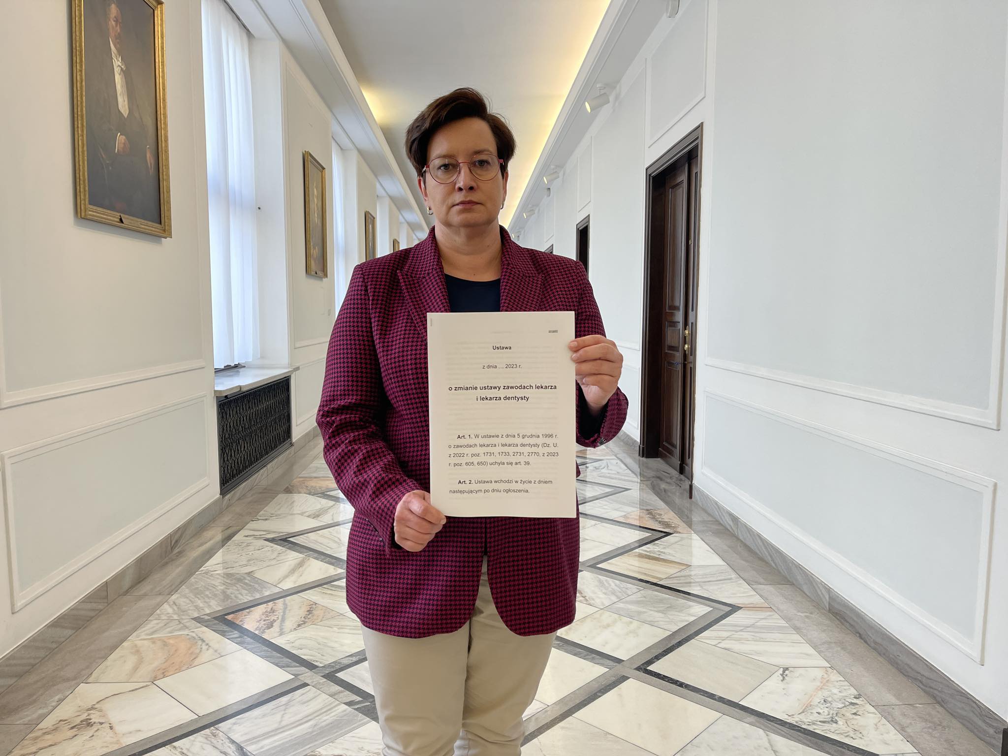 Kobieta w burgundowym żakiecie stoi w jasnym, urzędowym korytarzu i trzyma dokument z projektem uchwały znoszącej klauzulę sumienia.