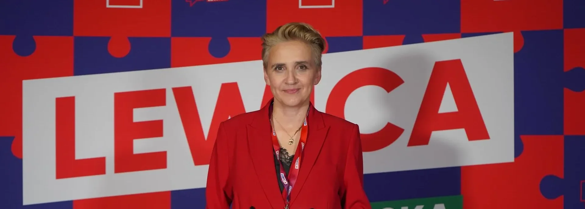 Uśmiechnięta kobieta w czerwonym garniturze stoi na tle kolorowych puzzli z dużym napisem LEWICA.