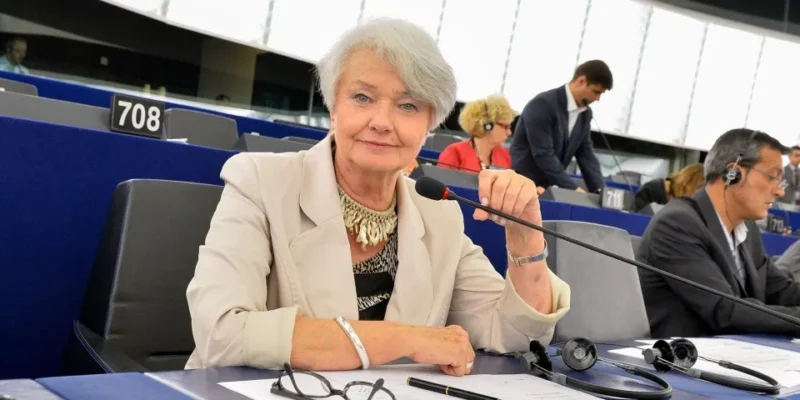 Kobieta o siwych włosach siedzi przy mównicy w sali obrad, trzyma mikrofon, a na biurku przed nią leżą dokumenty i słuchawki.