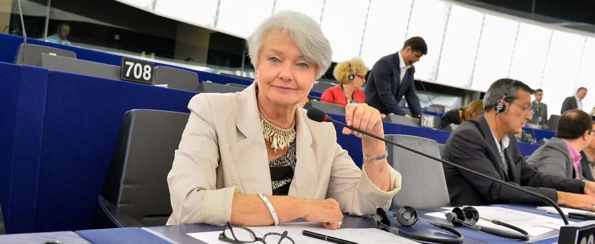 Kobieta o siwych włosach siedzi przy mównicy w sali obrad, trzyma mikrofon, a na biurku przed nią leżą dokumenty i słuchawki.