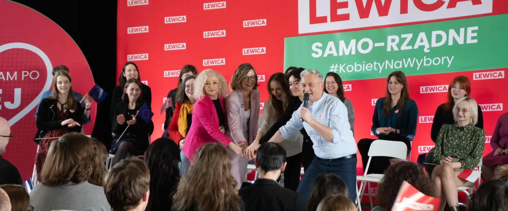 Kandydaci Lewicy na scenie wiecu wyborczego, witający wyborców i ściskający dłonie, z czerwonym tłem z logo LEWICA i zielonym banerem SAMO-RZĄDNE #KobietyNaWybory.