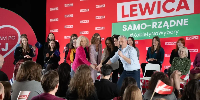 Kandydaci Lewicy na scenie wiecu wyborczego, witający wyborców i ściskający dłonie, z czerwonym tłem z logo LEWICA i zielonym banerem SAMO-RZĄDNE #KobietyNaWybory.