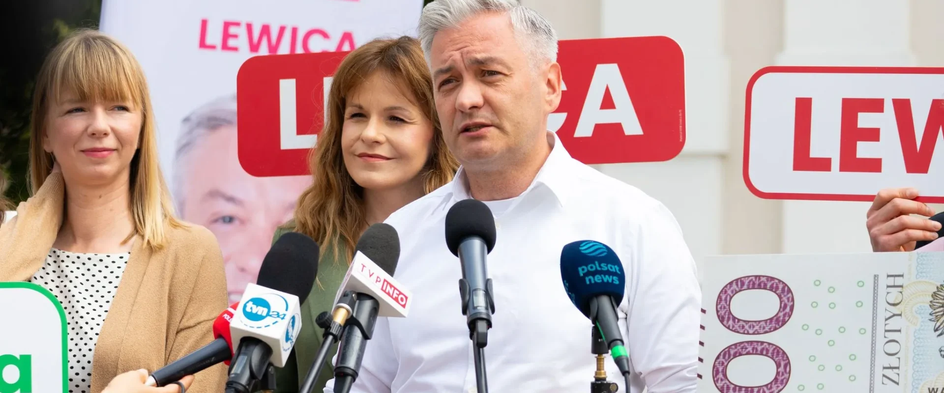 Polityk Lewicy przemawia na konferencji prasowej, stojąc przy mikrofonach, w towarzystwie dwóch kobiet, z tyłu widać banery z napisem LEWICA.