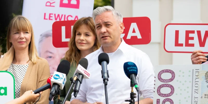 Polityk Lewicy przemawia na konferencji prasowej, stojąc przy mikrofonach, w towarzystwie dwóch kobiet, z tyłu widać banery z napisem LEWICA.