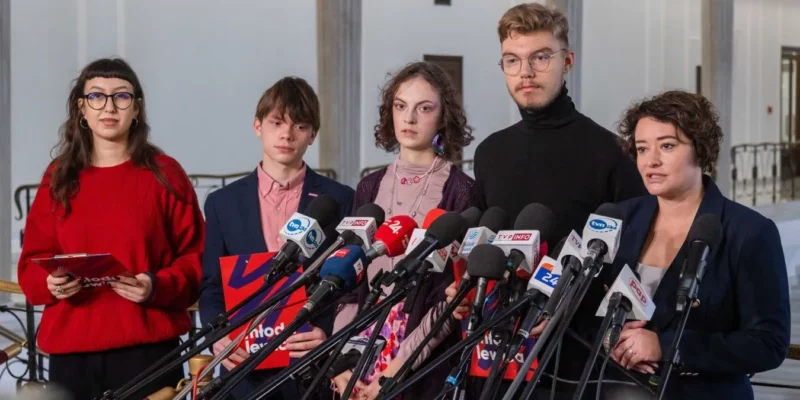 Grupa młodych ludzi stoi na konferencji prasowej przed licznym zestawem mikrofonów ustawionych na stojakach.