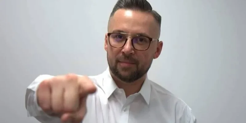 Mężczyzna w białej koszuli i okularach patrzy prosto w kamerę i wyciąga dłoń, wskazując palcem na widza.