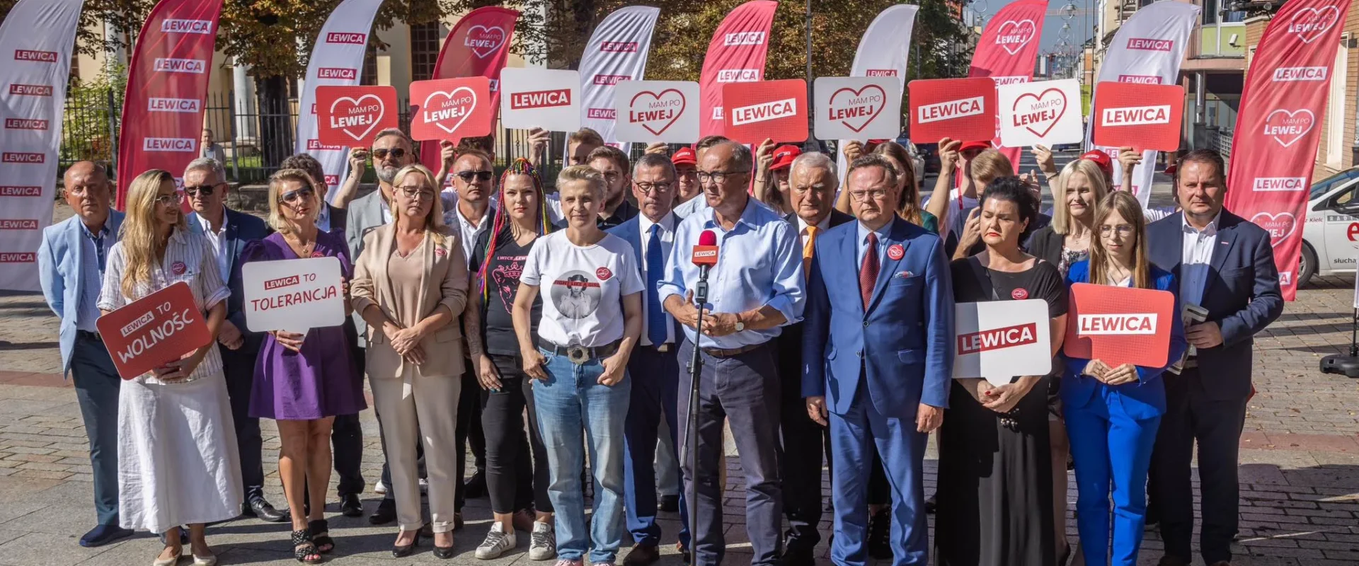 Grupa osób na placu trzyma plakaty i flagi z napisem LEWICA, widoczne hasła „LEWICA TO WOLNOŚĆ” i „LEWICA TO TOLERANCJA” oraz stoi przed mikrofonem.